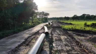 İşçiler, ağaçlar ve tarlalarla çevrili kırsal bir alanda bir gaz boru hattı inşa ederken görülüyor. Hazırlanan yol boyunca boruları yerleştirmek için ağır makineler kullanılıyor.