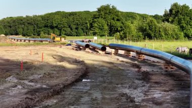 İnşaat faaliyetleri, kırsal bir bölgeye bir doğalgaz boru hattı kurulumunu gösteriyor. İşçiler bölgeyi hazırlarken ağır makineler çalışıyor. Yeşilliğin etrafını sarmak manzarayı güzelleştirir.