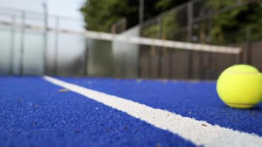 Oyuncular parlak mavi bir tenis kortunda antrenman yapıyorlar, sarı top yakınlarda dinlenirken yeteneklerini sergiliyorlar, servis edilmeye hazırlar. Ortam canlı ve enerjik..