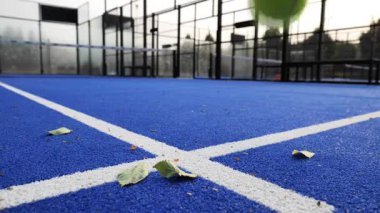 Canlı, mavi Padel kortu bir şehir parkında oynamaya hazır, yere saçılmış yapraklarla. Sabah ışığı yakındaki ağaçların arasından süzülüp davetkar bir atmosfer yaratıyor..