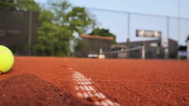 Tek bir tenis topu yerel bir spor tesisindeki kırmızı kil kortta nazikçe yuvarlanıyor. Çevre sakin, ağaçlar ve arka planda açık bir gökyüzü var..
