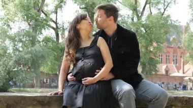 Bir çift, sıcak ve güneşli bir günde hamileliklerini kutlamak için yemyeşil bir parkta taştan bir bankta otururken şefkatli bir öpücük paylaşıyor. Mutlulukları ve aşkları doğada yayılıyor..