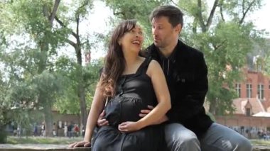 Bir koca ve hamile karısı huzurlu bir parkta samimi bir anı paylaşıyorlar. Birbirlerine gülümserler, güzelliğin ortasındaki ebeveynlik yolculuklarını kutlarlar..