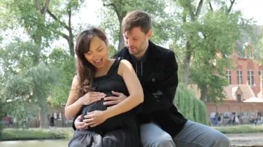 Bir çift, sakin bir park ortamında, hamileliği ve aşkı kutluyor. Güneş ışığı, gülümserken ve onun büyüyen karnını tutarken ılık bir parıltı yayar, bağlarını gösterir..