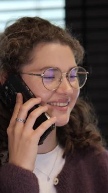 Kıvırcık saçlı ve gözlüklü genç bir kadın telefonunda neşeli bir sohbetten hoşlanıyor. Kapalı bir ortamda oturuyor, sıcaklığı ve dostluğu yayıyor..