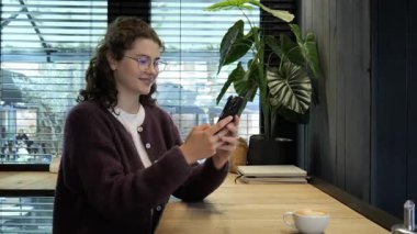 Sıcak ve davetkar bir yerde, genç bir kadın ahşap bir masada oturur, kahvesini yudumlar ve akıllı telefonunu kontrol eder. Atmosfer, dışarıda görünen yeşilliklerle rahatlıyor. Huzurlu bir zemin oluşturuyor..