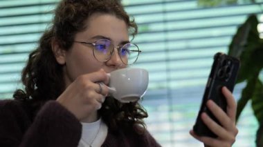 Genç bir kadın rahat bir kafede rahat rahat oturup sıcak bir fincan yudumluyor ve akıllı telefonuyla meşgul oluyor. Güneş ışığı pencereden süzülerek rahat bir atmosfer yaratır..