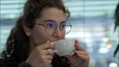 Bir kadın bir kafede rahatça oturup sıcak bir içeceğin tadını çıkarıyor. Doğal ışık pencerelerden süzülerek, rahatlaması için sıcak bir atmosfer yaratıyor..