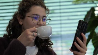 Kıvırcık saçlı ve gözlüklü genç bir kadın rahat bir kafede akıllı telefonuna bakarken fincandan kahve yudumluyor. Güneş ışığı pencereden süzülerek atmosferi güçlendirir..