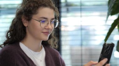 Kıvırcık saçlı ve gözlüklü genç bir kadın bir kafede akıllı telefonuna odaklanıyor. Güneş ışığı pencereden süzülerek sıcak ve davetkar bir atmosfer yaratır..