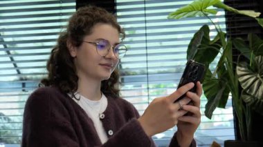 Genç bir kadın bir kafede rahat bir şekilde oturuyor, akıllı telefonuyla meşgul. Güneş ışığı pencereden süzülüp odaklanmış ifadesini ve etrafındaki yeşilliği vurguluyor..