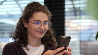 Kıvırcık saçlı ve gözlüklü genç bir kadın, sıcak, davetkar bir kafe ortamında akıllı telefonuyla konuşurken gülümsüyor. Yumuşak ışıklandırma gevşek atmosferi güçlendirir.