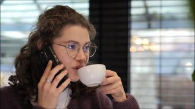 Kıvırcık saçlı genç bir kadın telefonda konuşurken kahvesini yudumluyor. Sıcak ışıklandırmalı rahat bir kafe ortamında neşeli ve rahat görünüyor..