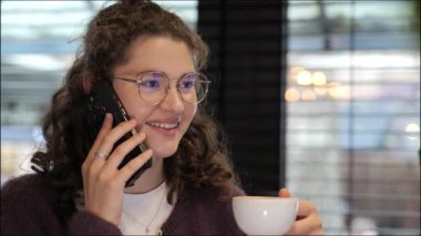 Kıvırcık saçlı genç bir kadın telefonuyla konuşurken gülümsüyor, sıcak, davetkar bir kafede elinde bir fincan kahve tutuyor. İçi hafif aydınlatma ve büyüleyici dekorlarla dolu..