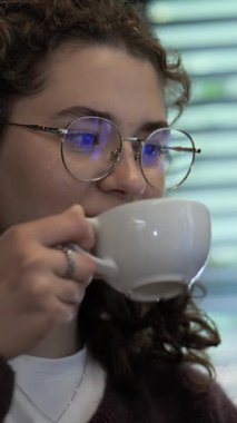 Genç bir kadın sakin bir ifadeyle bardaktan yudumluyor ve içkisinin tadını çıkarıyor. Yumuşak ışıklandırması ve sıcak atmosferi olan rahat bir kafede oturuyor. Rahatlamak için mükemmel..