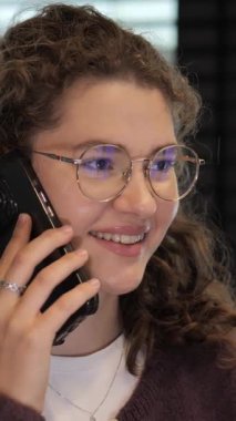 Genç bir kadın bir telefon görüşmesi yapar, neşeli bir şekilde gülümser ve rahat bir tavır sergiler. Yumuşak ışıklandırma onun coşkusunu ve modern çevresini vurguluyor..