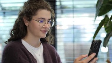 Genç bir kadın şık bir kapalı mekan ortamında akıllı telefonuna odaklanmış durumda. Perdelerden doğal ışık süzülüyor. Çevresinde meşgul ve rahat görünüyor..