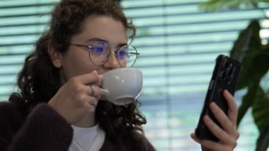 Genç bir kadın bir kafede rahatça oturup kahvesini yudumluyor ve akıllı telefon ekranıyla konuşurken gülümsüyor. Doğal ışık ve yeşillik hoş bir atmosfer yaratır..
