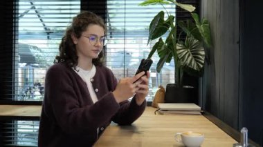 Genç bir kadın modern bir kafede ahşap bir masada oturuyor, kahvesini yudumluyor ve akıllı telefonuna odaklanıyor. Çevrede bitkiler ve geniş pencereler var..