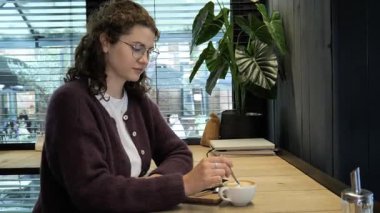 Kıvırcık saçlı bir kadın, rahat bir kafedeki ahşap bir masada oturup sıcak bir kahvenin tadını çıkarıyor. Ortam yemyeşil ve rahat bir ortama sahip..