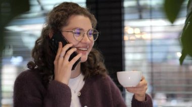 Kıvırcık saçlı ve gözlüklü genç bir kadın telefonda konuşurken gülümsüyor, elinde doğal ışık dolu sıcak ve davetkar bir kafe atmosferinde bir bardak tutuyor..