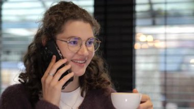 Kıvırcık saçlı ve gözlüklü genç bir kadın modern bir kafede kahve içerken telefonla konuşurken gülümsüyor. Atmosfer rahat ve davetkar. Sıradan sohbetler için mükemmel..