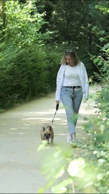 Bir kadın yemyeşil bir parkta köpeğiyle güneşli bir yol boyunca rahatça dolaşmaktan hoşlanır. Huzurlu ortam rahatlama ve bağlantı için mükemmel bir ortam yaratıyor..