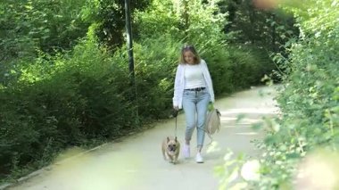Bir kadın, doğa yolunda yürürken köpeğine bağlı bir tasmayla huzurlu bir şekilde dolaşmaktan hoşlanır. Yeşillik onları çevrelemiş ve sakin bir atmosfer yaratmış..