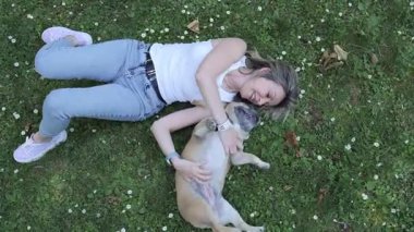 A woman lies on the grass in a park, smiling and laughing as she plays with her dog. The sun shines brightly, illuminating their joyful interaction amidst blooming flowers.
