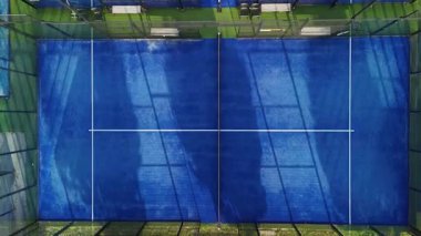 Bir raket sahası canlı mavi bir yüzeye sahip oyuncular için hazır bekliyor. Sahne, öğleden sonra güneş ışığını yakındaki bankları ve ekipmanları gösteren gölgeleri yakalıyor..