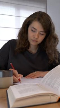 Genç bir kadın, iyi ışıklandırılmış bir masada sıcak bir içecek içerken ders kitabından notlar alarak derinden çalışmalarıyla meşgul. Atmosfer sakin ve yoğundur..