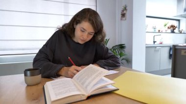 Genç bir insan, eğitici materyaller ve sıcak bir atmosferle çevrili, parlak bir mutfaktaki ahşap bir masada ders çalışmaya ve notlar yazmaya odaklanır..