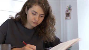 Kıvırcık saçlı genç bir kadın, iyi aydınlatılmış bir odada masasında özenle ders çalışıyor. Malzemelerine konsantre olurken notlar alıyor, eğitime olan bağlılığını gösteriyor..