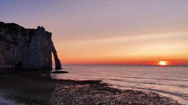 Güneş ufukta batarken Fransa 'daki Etretat uçurumlarının nefes kesici hava perspektifini görüntüler. Gökyüzünü canlı renklerle aydınlatır ve denizi yansıtır..