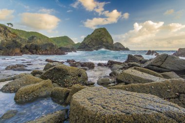 Okyanus Dalgaları Tropikal bir sahilde kayalara çarpıyor. Lush Green Hills ve Cloudy Sky Telawas Beach Lombok Endonezya 'ya doğru yavaşça süzülüyor.