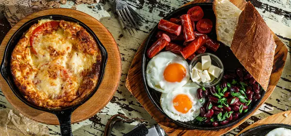 Set of delicious breakfasts, fried eggs with toasts and sausage on hot pan, english breakfast with beans and omelette with cheese on pan over wooden background. Healthy food, top view