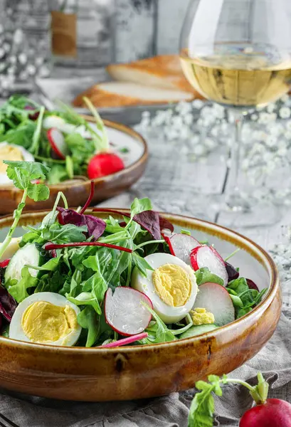 Summer fresh salad with eggs, radish, cucumbers and greens, arugula, leaves of lettuce in a plate on light wooden background served with glasses, wine, flowers. Healthy vegan food, clean eating