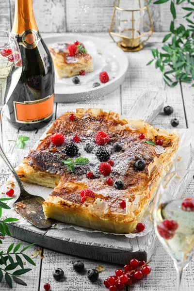 Holiday cheesecake with berries and powder sugar on light wooden background served with glasses, bottle of wine, candles, plants, festive decoration. Sweet dessert, close up