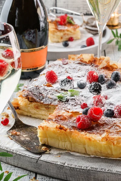 Holiday cheesecake with berries and powder sugar on light wooden background served with glasses, bottle of wine, candles, plants, festive decoration. Sweet dessert, close up