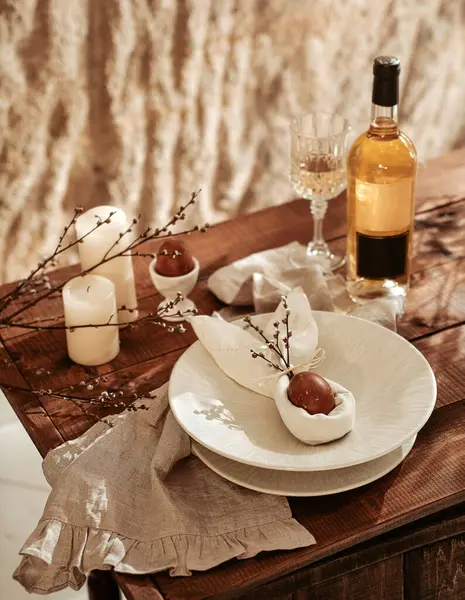 Easter holidays table, wine in glasses, painted eggs, served plates, willow branches, candles, bottle of wine on wooden background for festive dinner at home. Holiday concept