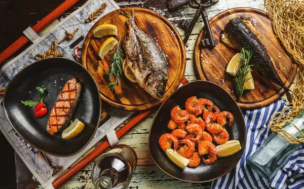 Seafood, grilled dorado fish, grilled salmon steak, smoked mackerel fish and fried shrimps with lemon and rosemary on wooden background with map and fishnet. Healthy food, top view