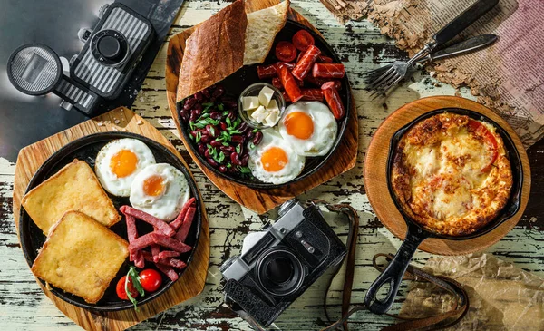 Set of delicious breakfasts, fried eggs with toasts and sausage on hot pan, english breakfast with beans and omelette with cheese on pan over wooden background. Healthy food, top view