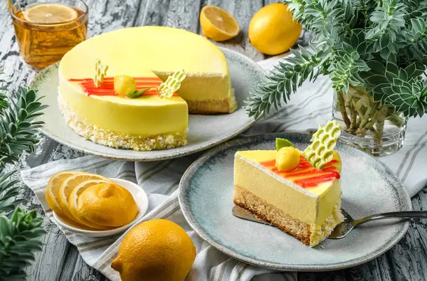Ayna jöleli sarı kremalı pasta, açık ahşap arka planda limonlar ve bir fincan çay ve bitkiler. Tatlılar, tatlı ve pasta, yakın çekim, seçici odaklanma