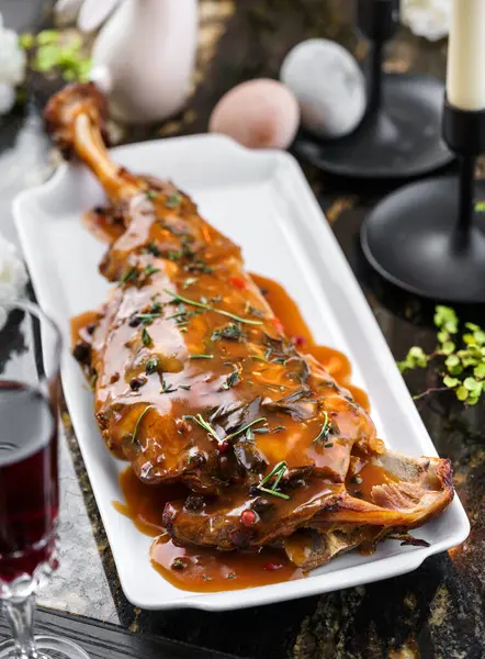 Baked lamb thigh leg, delicious meat for holiday with sauce and herbs on marble table with easter decoration and wine glasses. Happy Easter food, top view