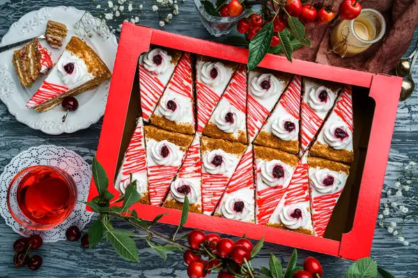 Pieces of delicious cake with cream and jam from cherry in box and on the plate over grey wooden background with fresh cherries on branch. Sweets, dessert and pastry, top view