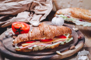 Çıtır çıtır ve lezzetli Fransız kruvasan sandviçi jambon, pastırma, peynir, marul, ahşap kesim tahtasında domatesler. Et, kahvaltı, hamur işi, yakın çekim.
