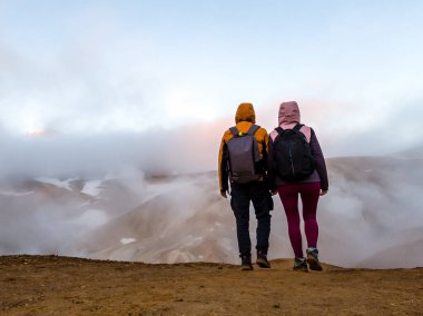 Turuncu tepenin tepesinde sırt çantalı iki turist dağ sırasının geri kalanının manzarasına bakıyor. Arka planda jeotermal alandan gelen buhar. Turistler sarı ve pembe ceket giyiyorlar ve başlıkları da var..