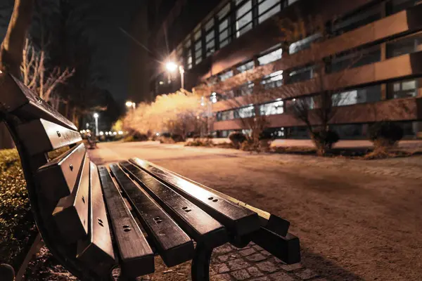 Şehir parkındaki boş ahşap bankın kapağını kapat. Arka planda patika ve modern bina var. Görünürde kimse yok.