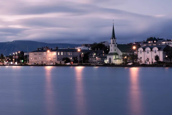 Reykjavik şehir merkezinin Tjrnin Gölü üzerindeki Frkirkjan kilisesi ile akşam manzarası gökyüzünde bulanık bulutlar ve ön plandaki suya yansıyan ışık. 