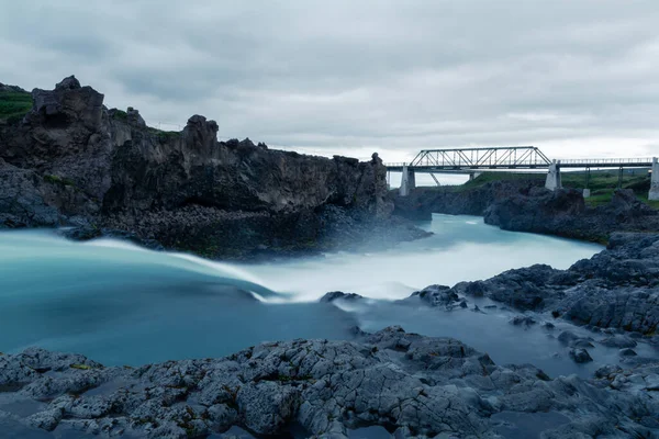 蓝水瀑布的景观从两侧锋利的黑暗岩石上滑落下来 混凝土柱子上的钢桥背景 多云的天空 长期曝光照片 — 图库照片